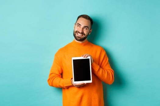 Technology. Handsome adult man showing digital tablet screen, smiling and looking at upper left corner thoughtful, imaging or daydreaming, blue background.
