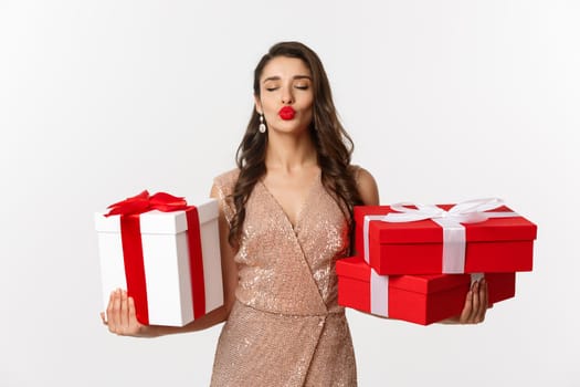 Holidays, celebration concept. Beautiful caucasian woman in elegant dress holding Christmas presents and smiling happy, standing over white background.