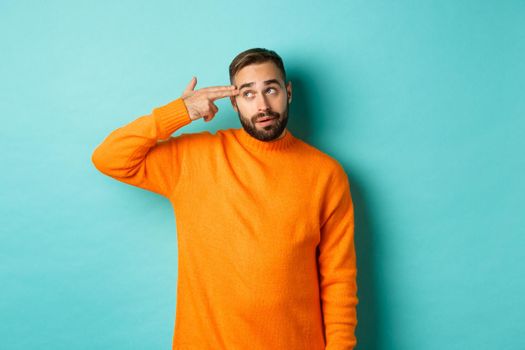 Man shooting himself in head, make finger gun near temple and roll eyes annoyed, standing bothered against light blue background.