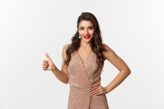 Concept of celebration, holidays and party. Attractive and confident woman with red lips, wearing luxury evening dress and showing thumb up, approve and like, white background.