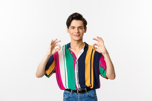 Satisfied young queer man showing okay signs and smiling in approval, agree and like something, praise excellent choice, standing over white background.