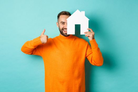 Real estate concept. Satisfied man recommending agency, showing thumb up and house maket, standing over blue background.