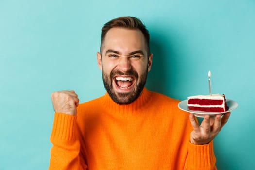 Close-up of happy adult man celebrating birthday, holding bday cake with candle and making wish, standing against turquoise background.