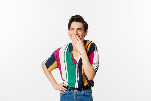 Happy young man laughing and covering mouth with hand, looking up at something funny, standing over white background.