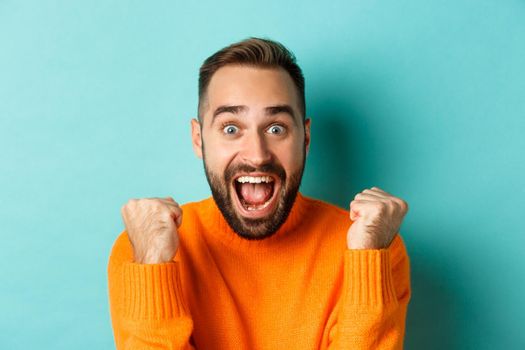 Image of handsome relieved man feeling satisfaction, rejoicing of winning or achievement, making fist pump and saying yes, standing over turquoise background.
