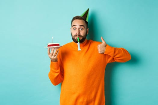 Happy birthday guy celebrating, wearing party hat, blowing wistle and holding bday cake and showing thumb-up in approval, standing over turquoise background.