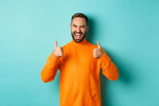 Super good. Satisfied happy man showing thumbs up, agree with you, praise excellent work, looking pleased, standing in orange sweater against light blue background.