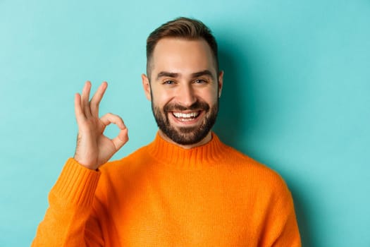 Close-up of assertive young man assuring everything ok, showing okay sign and smiling, yes or positive answer, standing over light blue background.