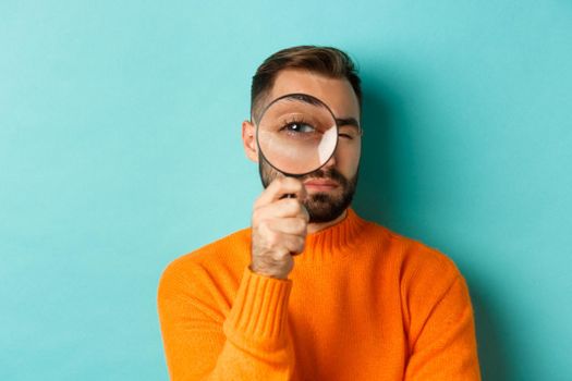Funny man looking through magnifying glass, searching or investigating something, standing in orange sweater against turquoise background.