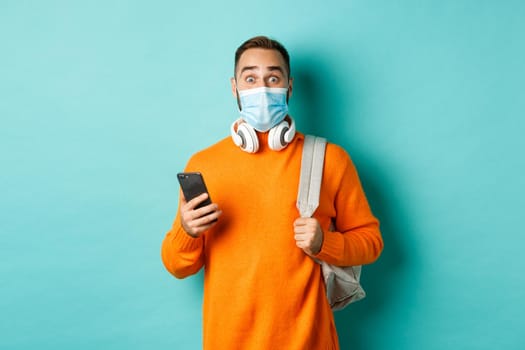 Young man in face mask using mobile phone, holding backpack, staring impressed at camera, standing against light blue background.