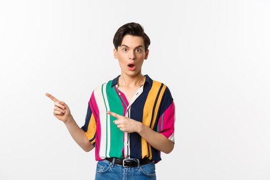 Amazed young queer man pointing fingers left, staring at camera with disbelief, asking question about product, standing over white background.