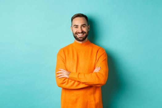 Handsome bearded male model in orange sweater, looking confident and smiling, cross arms on chest, standing over turquoise background.