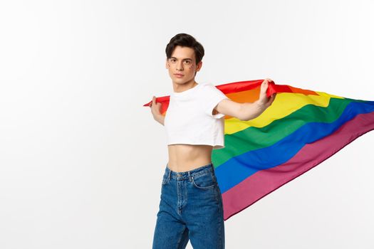 Human rights and lgbtq community concept. Out and proud gay man waving rainbow flag and looking confident at camera, standing in crop top and jeans against white background.
