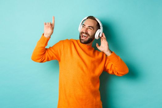 Happy adult man in orange sweater, looking up and listening music in headphones, standing over blue background.