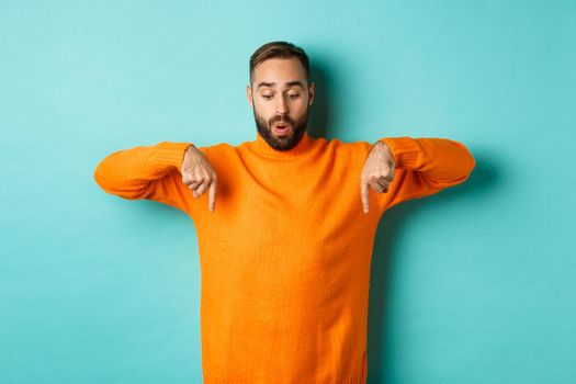 Amazed guy in orange sweater, pointing fingers down and looking with interest at promo, holiday discounts, standing over turquoise background.