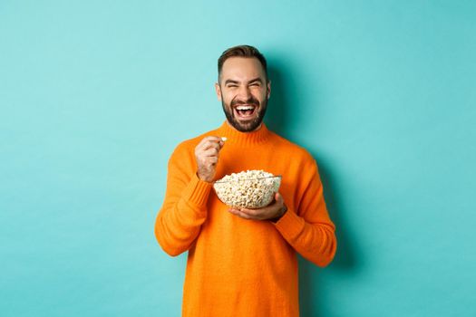 Excited young man watching interesting movie on tv screen, eating popcorn and looking amazed, blue background.