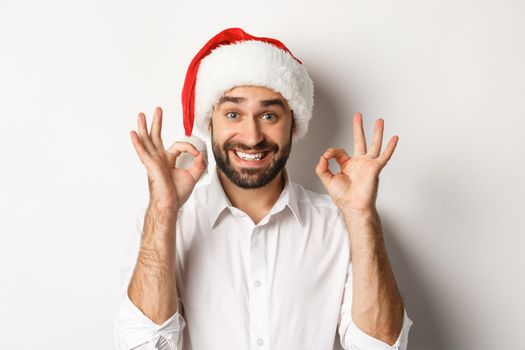 Party, winter holidays and celebration concept. Joyful man enjoying christmas and showing okay sign, smiling satisfied, wearing santa hat, white background.