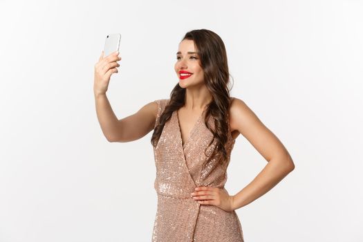 Beautiful woman with makeup and hairstyle, wearing glamour dress, taking selfie on smartphone, celebrating christmas party, white background.