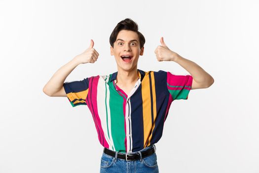 Excited and amazed young man showing thumbs up with impressed expression, standing over white background.