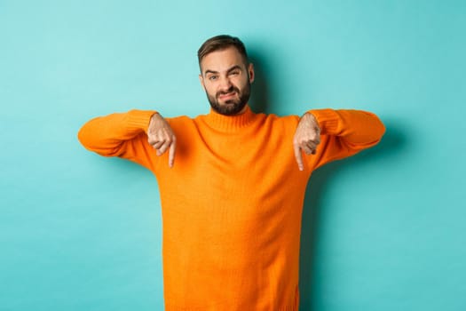Displeased and skeptical handsome man pointing fingers down, showing bad product, dislike store or advertisement, grimacing disappointed, standing over turquoise background.