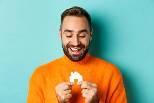 Real estate concept. Happy young man searching for home rent, holding house paper maket and smiling, standing over blue background.