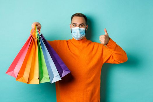 Covid-19, social distancing and lifestyle concept. Young man in face mask showing shopping bags and thumb-up, buying holiday gifts during pandemic.