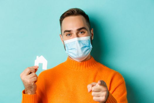 Real estate and coronavirus pandemic concept. Close-up of adult man in medical mask pointing at you and showing small paper house, standing over light blue background.
