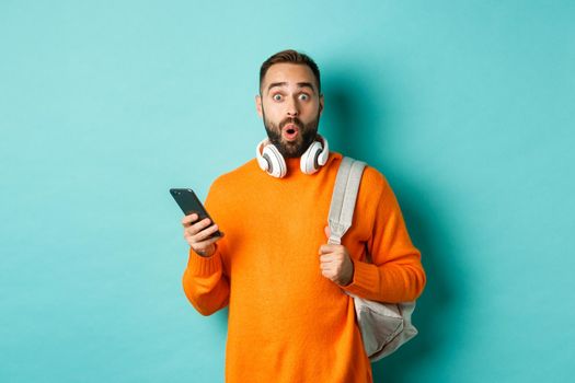 Caucasian man with headphones and backpack staring at camera surprised after reading phone message, standing over turquoise background.
