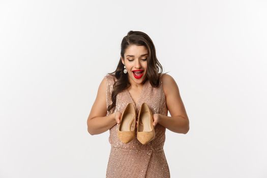 Party and celebration concept. Excited woman showing new pair of heels and smiling, wearing elegant dress, standing over white background.