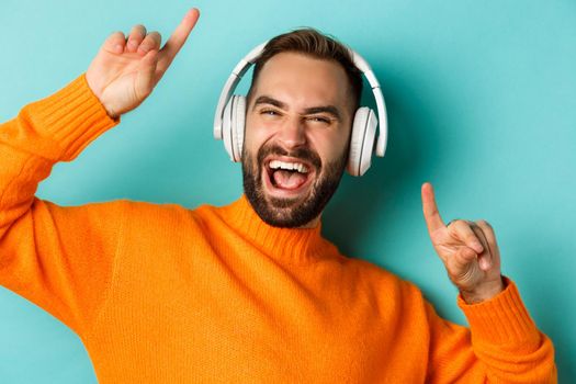 Close-up of handsome modern man listening music in headphones, standing in orange sweater over turquoise background.