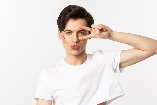 People, lgbtq community and lifestyle concept. Close-up of sassy and beautiful gay man with glitter on face, pucker lips in kiss and showing peace sign, white background.