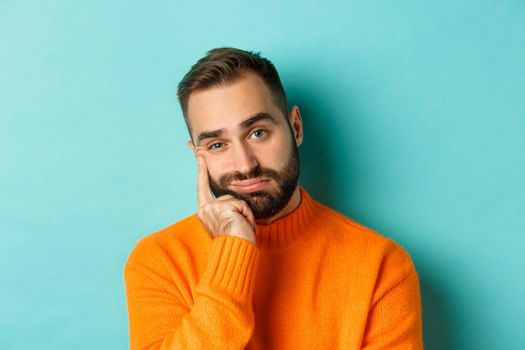 Close-up of unamused sad man looking at camera, pouting and leaning face on hand, standing over light blue background.
