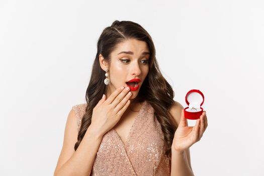 Surprised beautiful woman looking at engagement ring, feeling touched, receive marriage proposal, standing over white background.