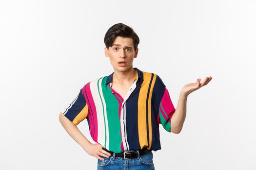 Confused gay man asking what happened, raising hand aside and looking puzzled at camera, standing over white background.