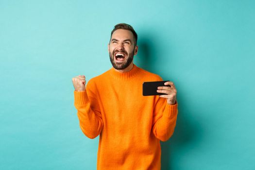 Image of man winning on mobile phone, making fist pump and rejoicing of victory, holding smartphone with satisfied face, turquoise background.