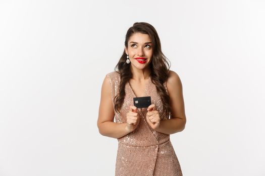 Holidays. christmas party and shopping concept. Beautiful female model in luxury dress thinking and holding credit card, standing over white background.