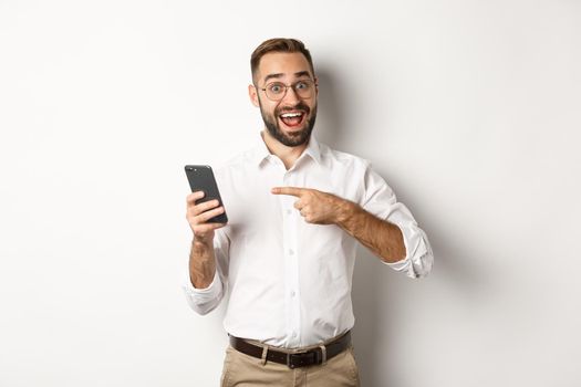 Man looking excited and pointing finger at mobile phone, showing good online offer, standing over white background.