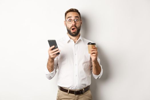 Image of handsome manage drinking coffee, reacting surprised to message on mobile phone, standing over white background.