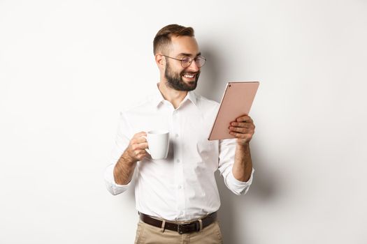 Handsome businessman drinking coffee and reading on digital tablet, smiling pleased, standing over white background.