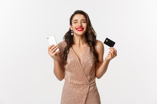 Online shopping and holidays concept. Satisfied and happy woman in elegant dress smiling, using credit card and mobile phone, standing over white background.