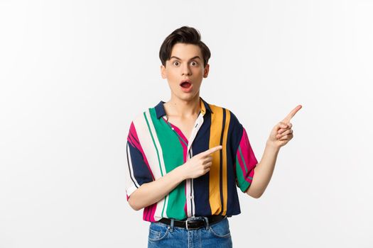 Impressed young man showing advertisement, open mouth with disbelief and pointing at upper right corner, standing against white background.