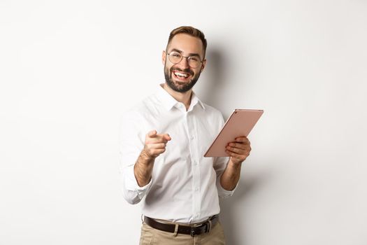 Happy and satisfied boss praising good job, reading on digital tablet and pointing at you camera, standing over white background.