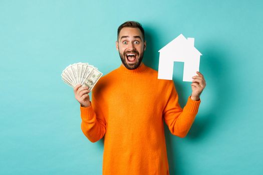 Real estate concept and mortgage concept. Excited man showing dollars and paper maket of house, buying flat, standing over blue background.