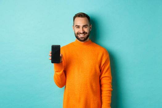 Handsome bearded guy in orange sweater, showing smartphone screen and smiling, showing promo online, turquoise background.