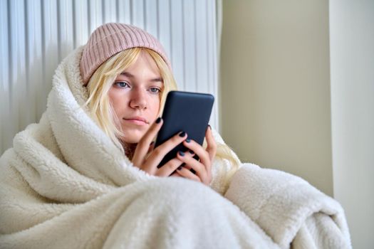 The cold season, cold winter and autumn, a frozen female teenager in a hat under a blanket sitting near a heating radiator with a smartphone in her hands