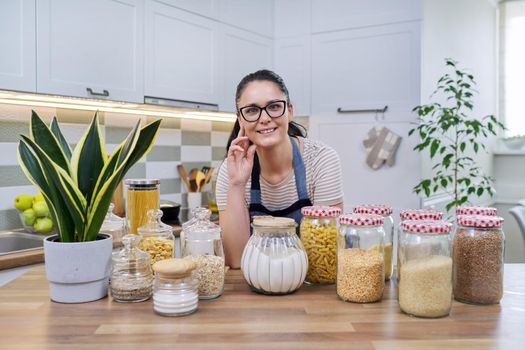 Food, grocery storage, smiling woman looking at the camera in the kitchen. Organization of storage of bulk products in glass jars and containers, pantry