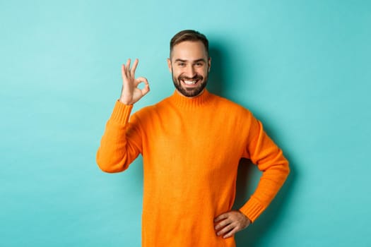 Image of confident handsome man showing ok sign, approve and agree, like something good, standing in orange sweater.
