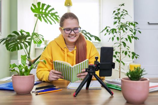 Teenage girl looking at webcam using smartphone for an online lesson, reading textbook book. Student 12, 13 years old, studying remotely at home, e-learning, virtual lessons, distance learning