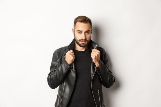 Image of handsome and confident man putting on leather biker jacket, standing against white background.
