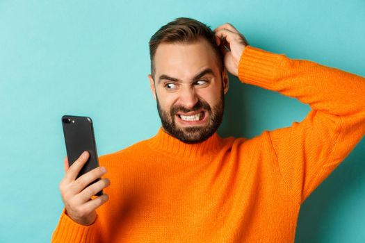 Close-up of caucasian man looking troubled and reluctant at phone screen, scratching head and grimacing confused, standing over turquoise background.